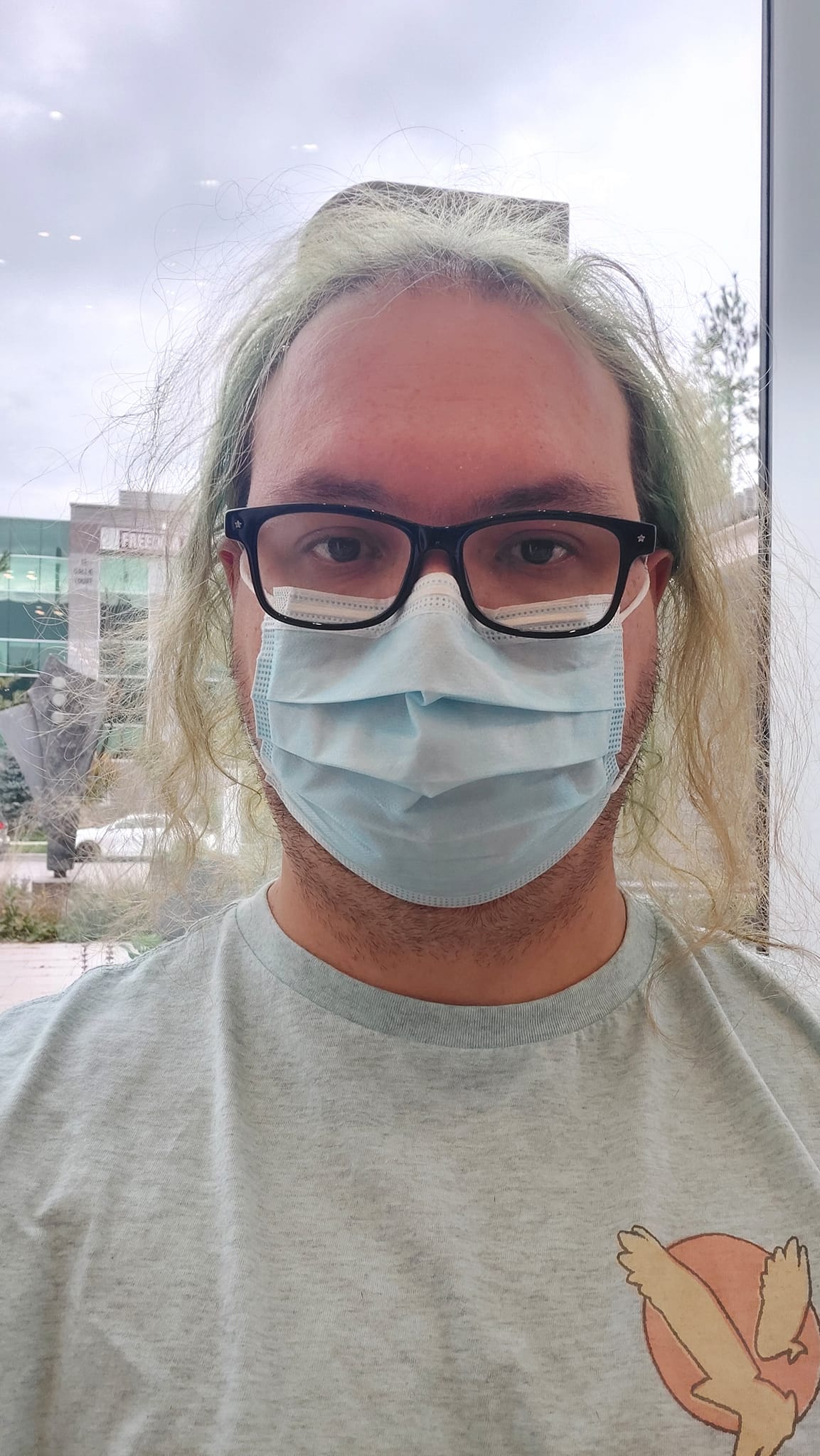 A man with long green hair wearing a light blue shirt and surgical mask sits next to a window in a hospital waiting room